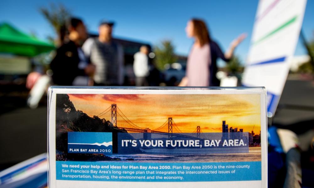 A tabletop sign reading "It's Your Future, Bay Area" with the Plan Bay Area 2050 logo is in the foreground while a staff member speaks with members of the public in the background.