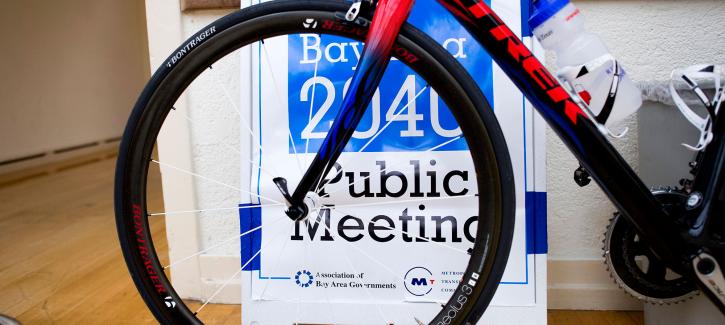 A Plan Bay Area 2040 Public Meeting sign is visible through the spokes of a bicycle tire.
